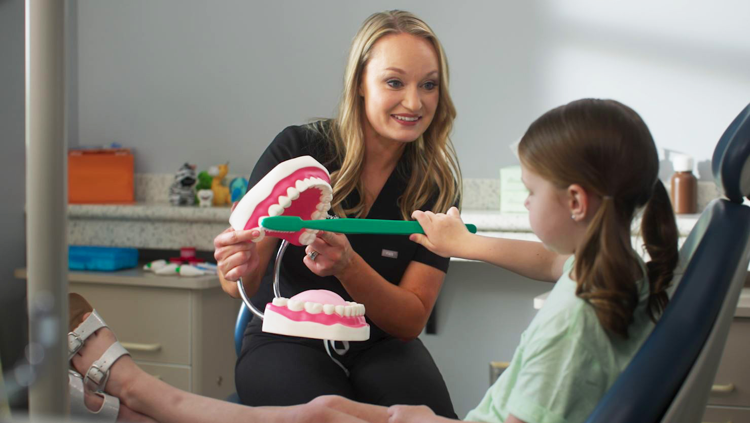 dr. bailey sits with her young patient brushing an oversized display set of teeth