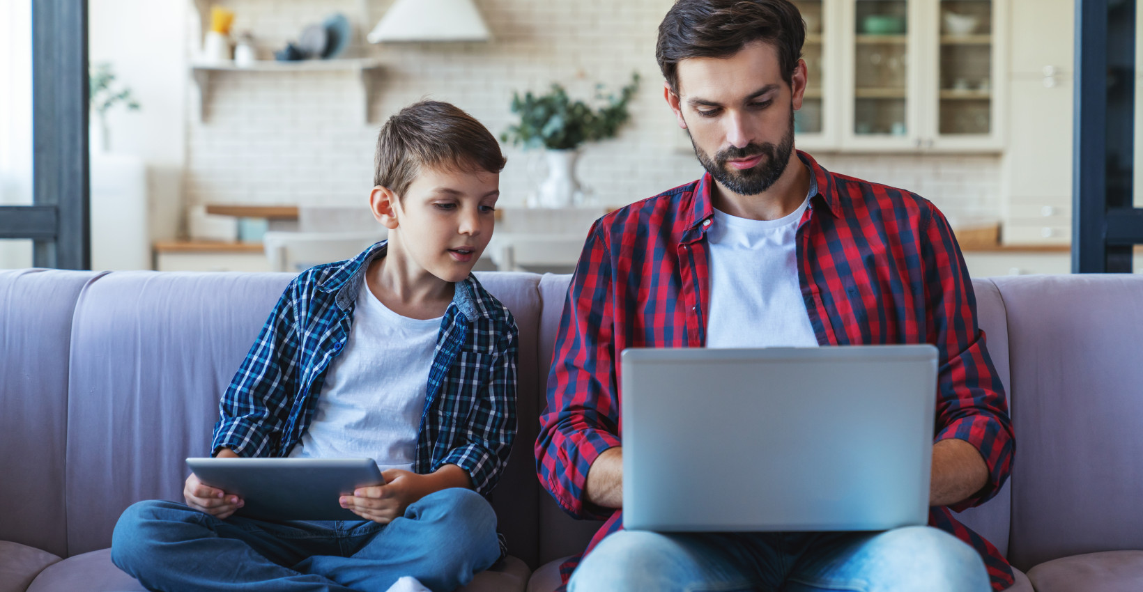 a father and son sit together on a couch looking at a laptop