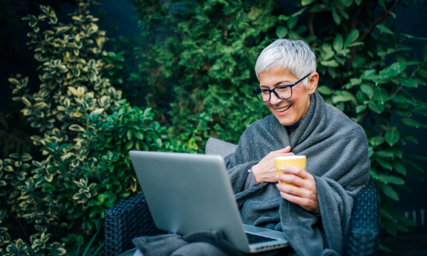 An elderly woman looking at a laptop and wrapping a blanket around herself. 