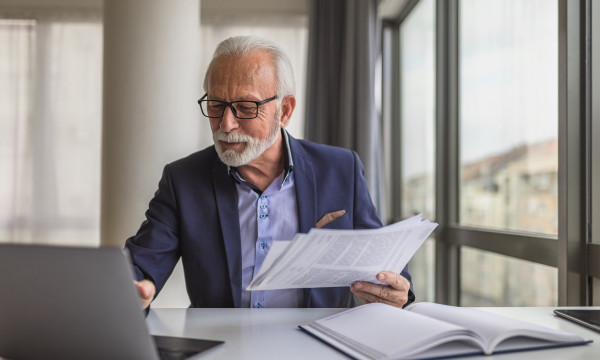 an older man wearing a blue suit sits at a computer with papers in his hands